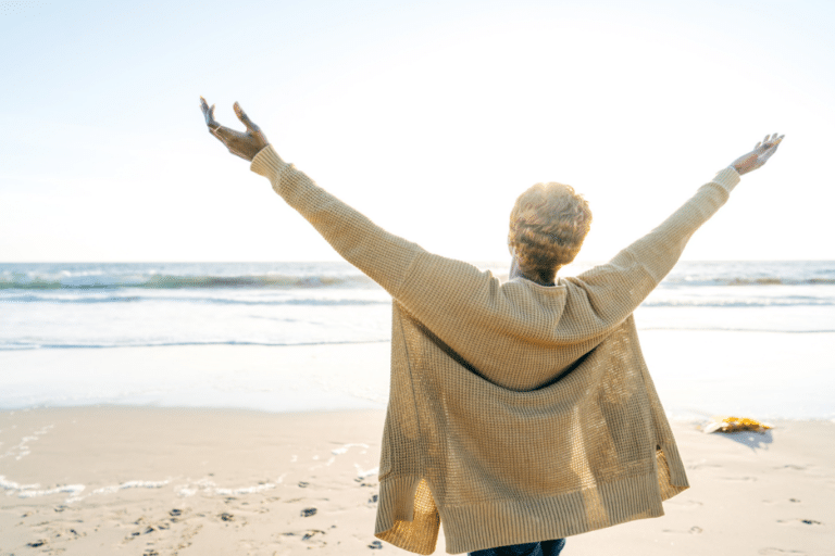 A woman on the beach facing the ocean raising her hands in the air.