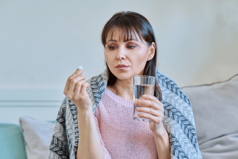 mature woman taking pill with glass of water, sitting under blanket at home