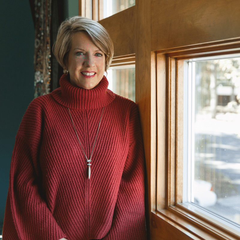 Dr. Anna Garrett, women's hormone specialist, smiling in red turtleneck sweater by window - warm and approachable menopause expert