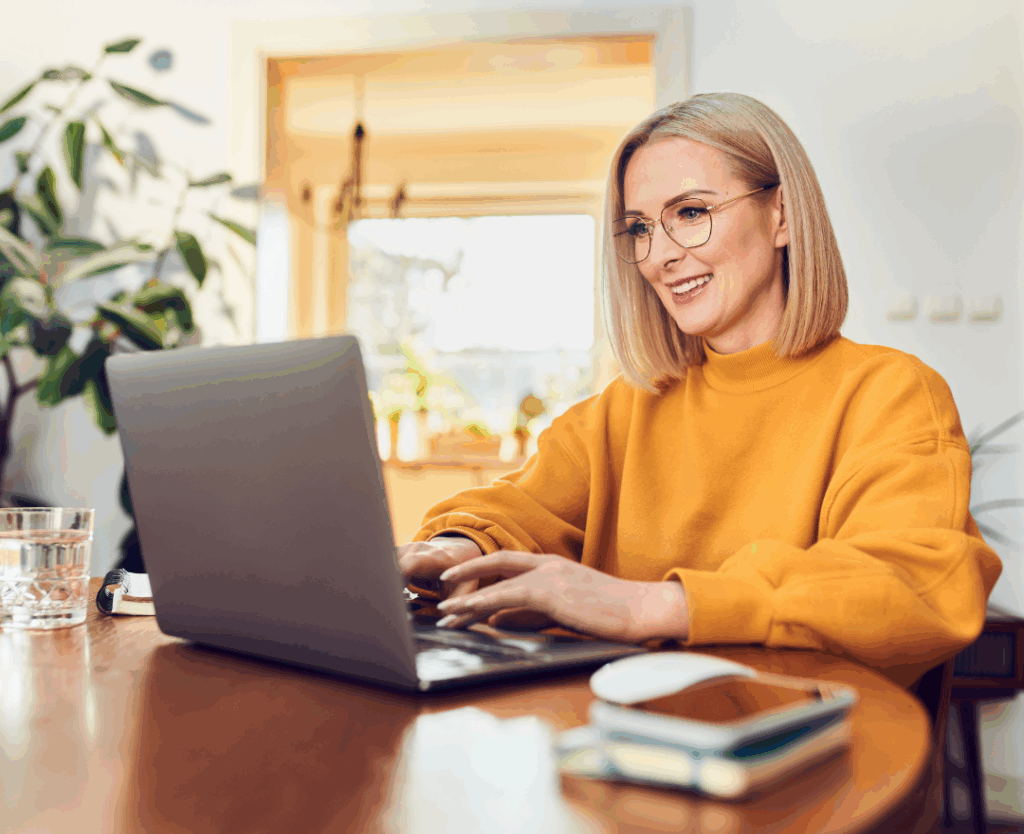 Smiling midlife blonde woman in yellow sweater working on laptop at home office desk - representing women taking control of their health