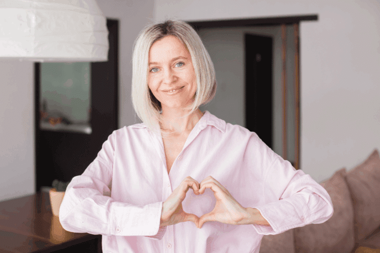 Smiling blonde woman in pink shirt making heart shape with hands, representing heart health and cardiovascular wellness for women in menopause