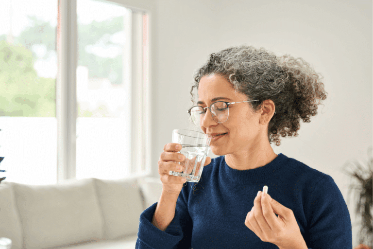 Happy healthy middle age woman taking supplements holding glass of water standing at home.
