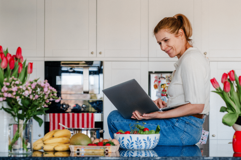 Midlife woman sitting on kitchen counter with laptop researching functional health coaching for perimenopause symptoms and fatigue