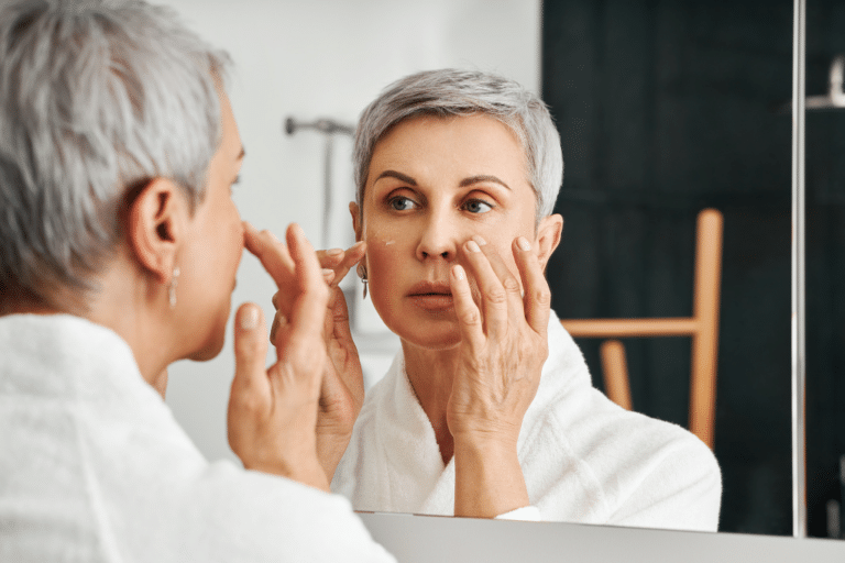 Midlife woman applying topical estrogen face cream while examining skin aging in bathroom mirror during menopause