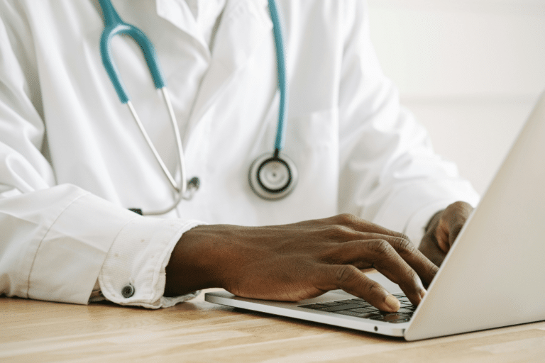 Doctor in white coat with stethoscope typing on laptop—reviewing annual hormone testing labs for postmenopausal women on estrogen and progesterone