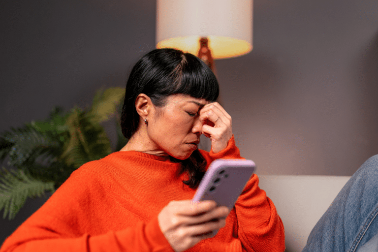 Overwhelmed midlife woman in an orange sweater pinching the bridge of her nose while reading messages on her phone, illustrating how to help someone grieving without adding to their load
