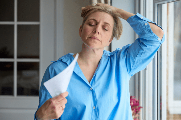 Midlife woman with eyes closed fanning herself with paper during a hot flash, a symptom treated with hormone therapy