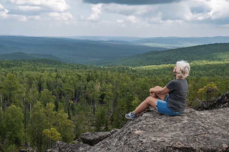 Midlife woman with gray hair sitting calmly on a rocky cliff edge, looking out over a green forest valley, a hopeful take on the menopause “physiological cliff” study